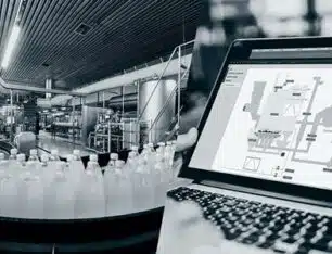 Teollisuusympäristö, jossa näkyy tuotantolinja ja kannettava tietokone, jonka näytöllä näkyy tuotantoympäristöä havainnollistava ohjelmisto